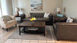 A living room with a grey sofa, a brown leather armchair, a coffee table with a vase of flowers, and a painting on the wall. - Surfside Shangri-La (Daytona Beach)