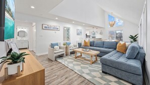 A spacious living room with a large grey sectional sofa, a wooden coffee table, and a checkered rug. There is a white reception desk with a plant, and a sign on the wall that reads "The Dream Beach House." - 2 blocks 2 beach, Golf Cart, Outdoor Bar, Dogs Welcome, Beach Gear (Atlantic Beach)