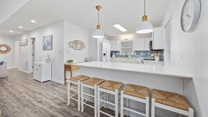 A modern kitchen with white cabinetry, a central island with bar stools, pendant lighting, and a clock on the wall. - 2 blocks 2 beach, Golf Cart, Outdoor Bar, Dogs Welcome, Beach Gear (Atlantic Beach)