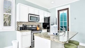 A kitchen with white cabinets, a microwave, a stove, and a refrigerator. There is a small island with a sink and bar stools. - Adorable Starlight EscapeCottage (Panama City Beach)