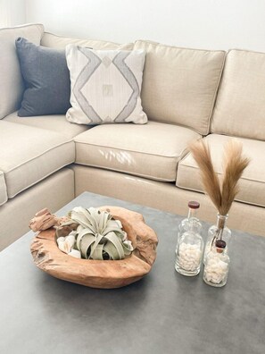 A beige sofa with a geometric patterned pillow, a wooden bowl with dried plants, and two glass vases with white pebbles and dried pampas grass on a grey table. - Moonlight and Starlight Escape (Panama City Beach)