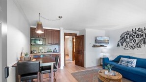 Una cocina moderna con armarios de madera y un comedor. Hay un sofá azul y una alfombra en la sala de estar. Un letrero de bienvenida está en la pared.
