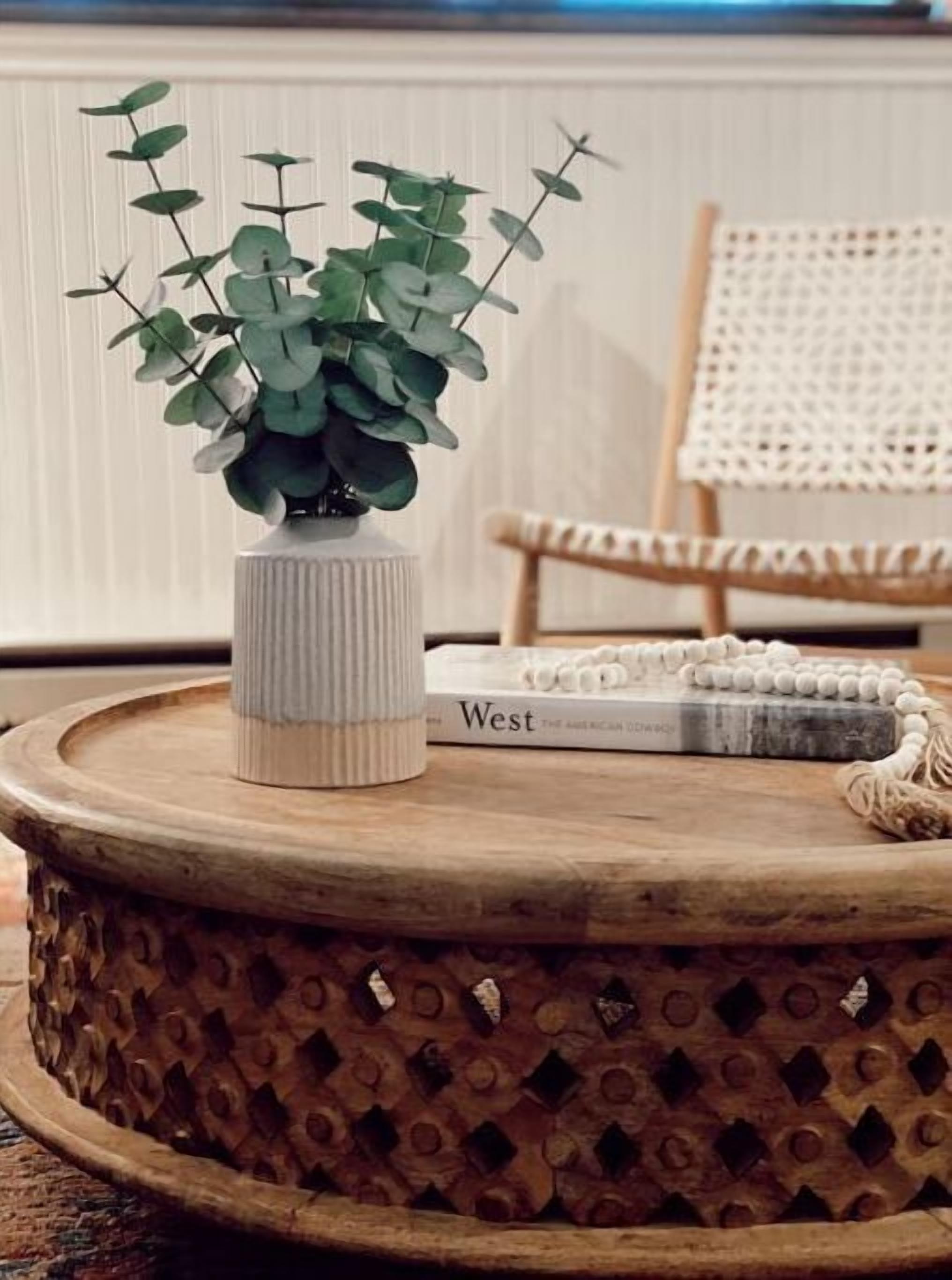 A round wooden table with a textured surface, a white vase with green foliage, and a book titled 'West'.