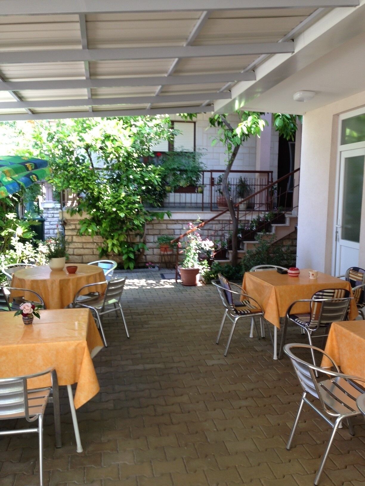 Un patio al aire libre con mesas y sillas, rodeado de plantas en macetas y una escalera que lleva a un nivel superior.