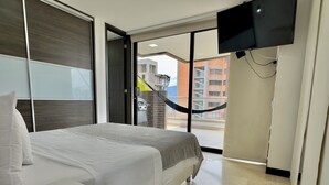 Una camera d'albergo con un letto, un comodino bianco, una TV montata a parete e vista su un balcone esterno con un'amaca.