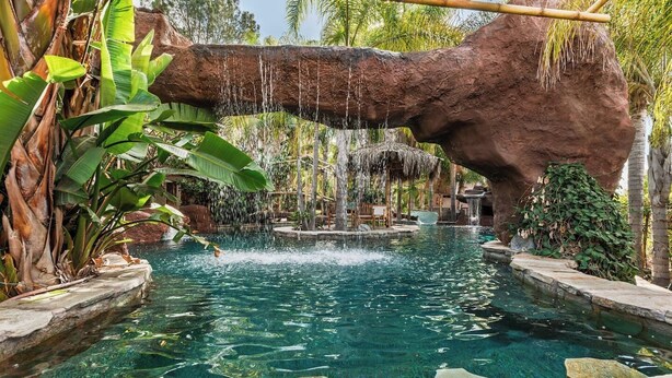 En klar pool med ett vattenfall, omgiven av tropiska växter och en stor stenformation.