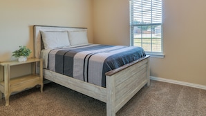 Een slaapkamer met een houten bed, een nachtkastje, een raam met jaloezieën en een vloerbedekking.