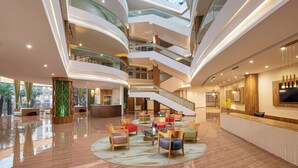 Un vestíbulo de hotel de varios niveles con una escalera central, zonas de estar y una mesa de recepción.