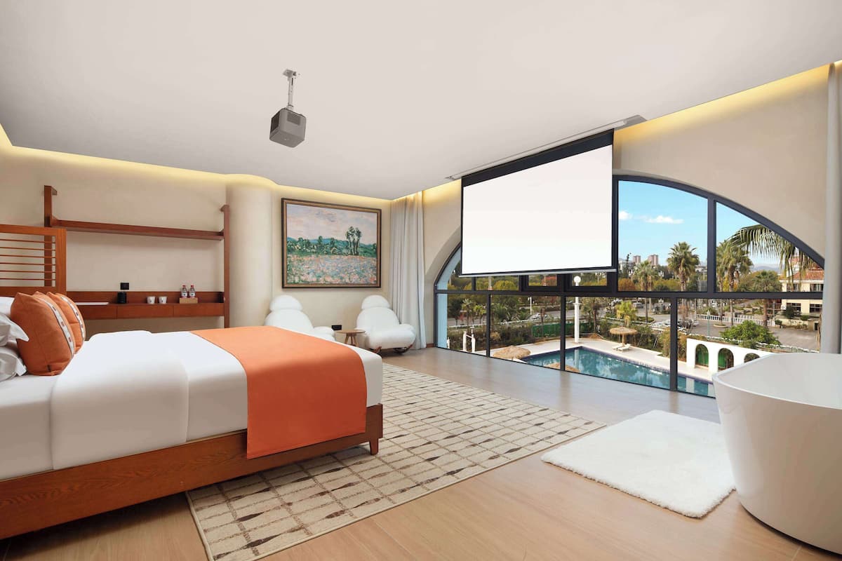 Une chambre d’hôtel moderne dotée d’un grand lit, d’un bureau, d’une chaise, d’une grande fenêtre donnant sur la piscine et d’un écran de projection.