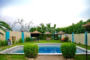 A swimming pool with a tiled border, surrounded by a well-maintained lawn and several thatched-roof structures.