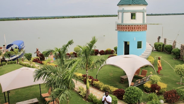 A tower with a blue and white exterior, surrounded by a well-maintained garden with palm trees and shrubs, and a body of water in the background.