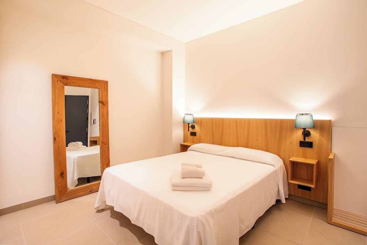 A hotel room with a wooden headboard, a bed with white linens, two wall-mounted lamps, and a wooden nightstand.