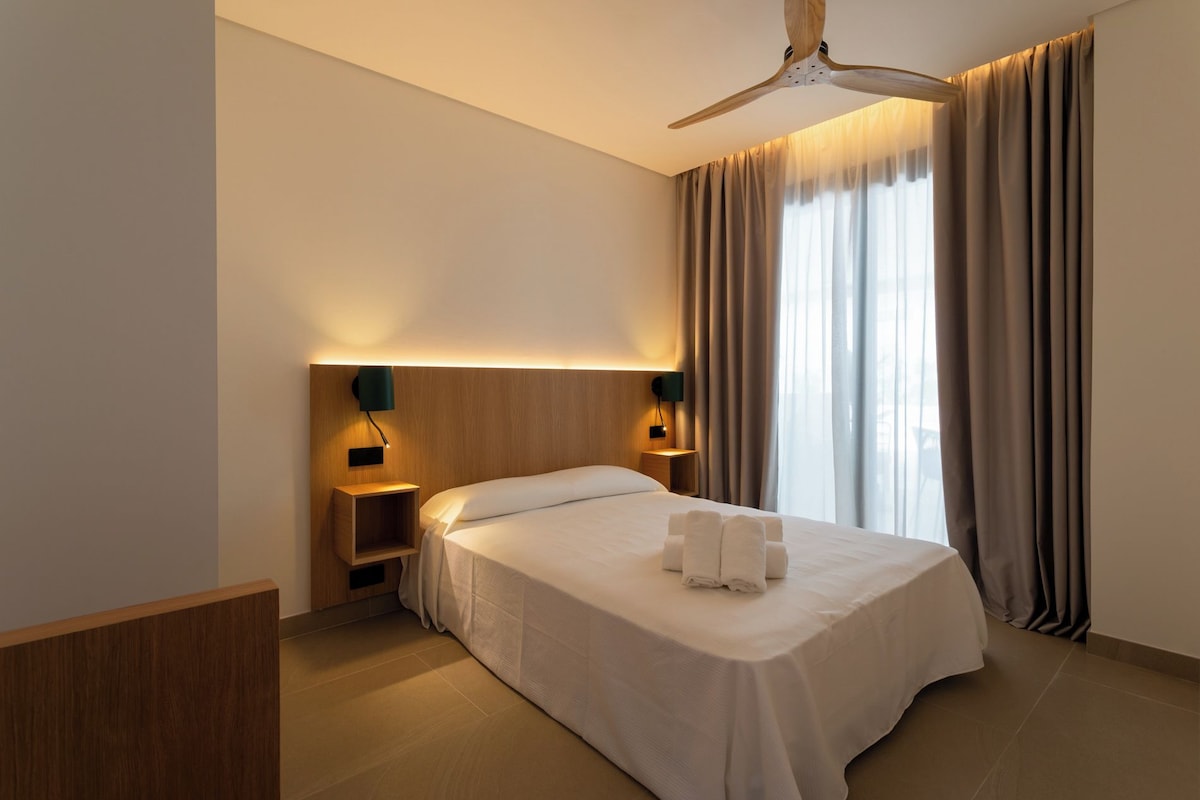 A hotel room with a bed, a ceiling fan, two bedside tables with lamps, a nightstand, and a window with curtains.
