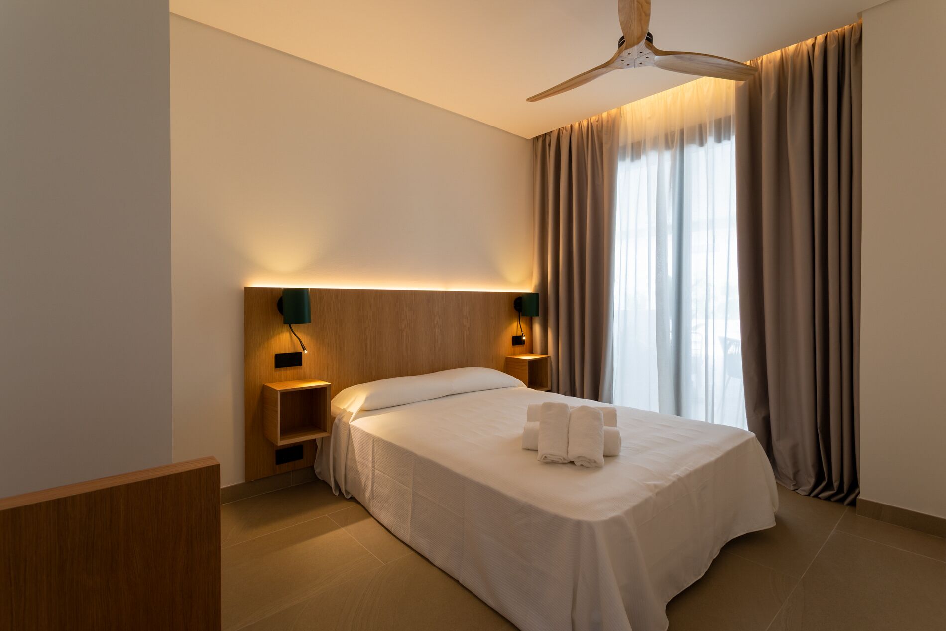 A hotel room with a bed, a ceiling fan, two bedside tables with lamps, a nightstand, and a window with curtains.