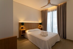 A hotel room with a bed, a ceiling fan, two bedside tables with lamps, a nightstand, and a window with curtains. - Hotel Apartamentos Hamus  (Conil de la Frontera)