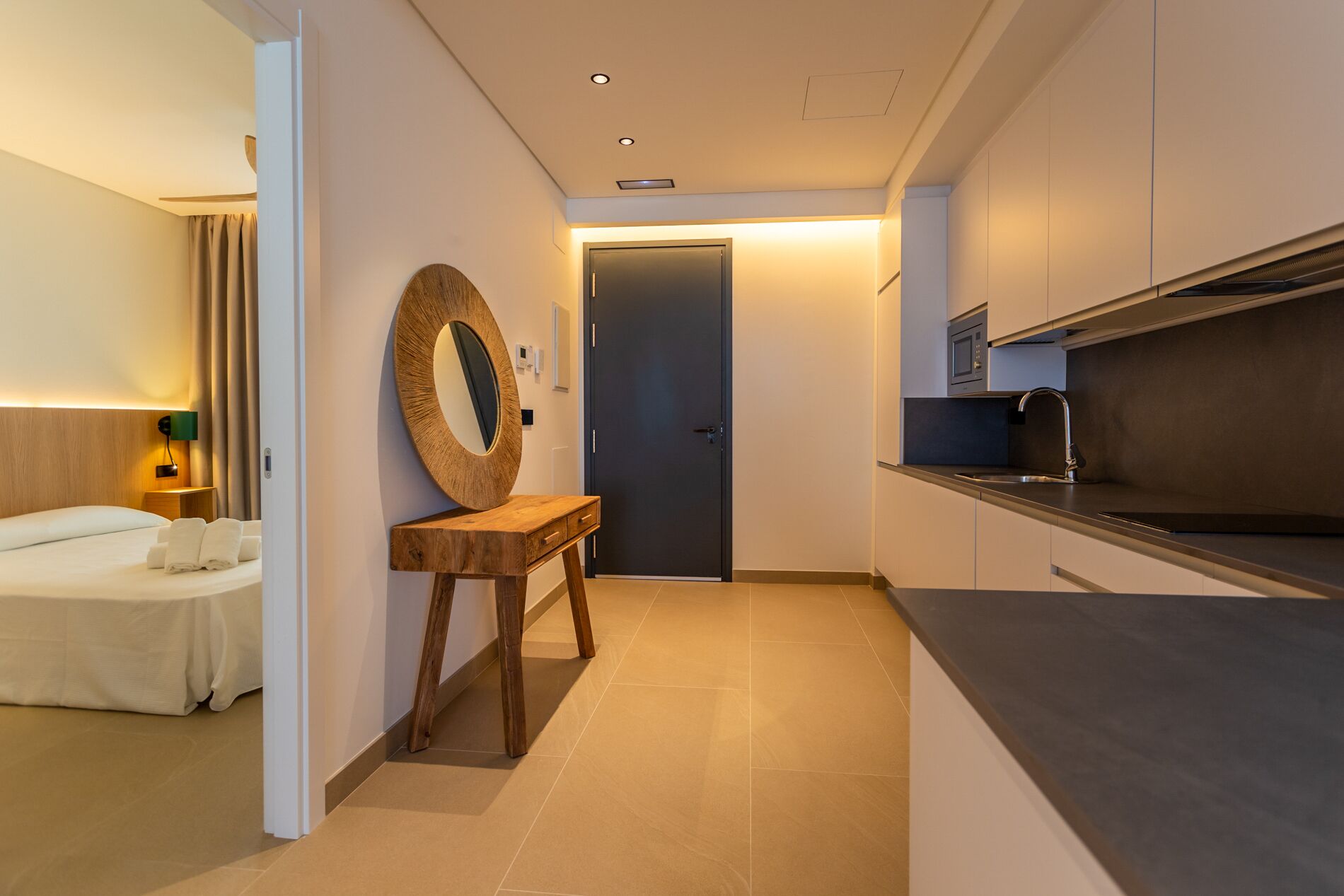 Una habitación de hotel moderna con una cama, una mesita de noche de madera con espejo, una puerta oscura y una kitchenette con microondas y fregadero.