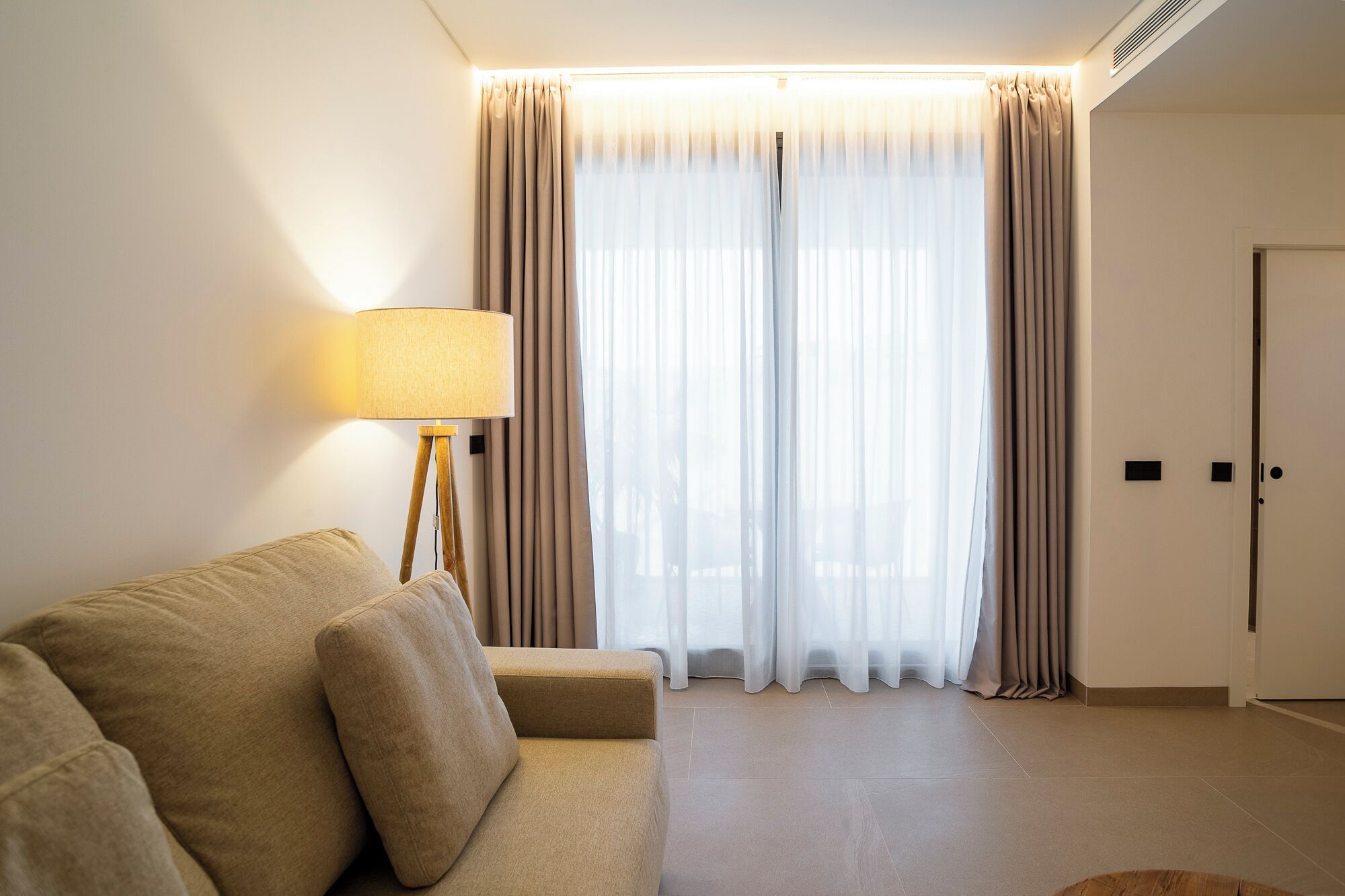 Una sala de estar moderna con un sofá color beige, una lámpara de pie y cortinas de gasa.