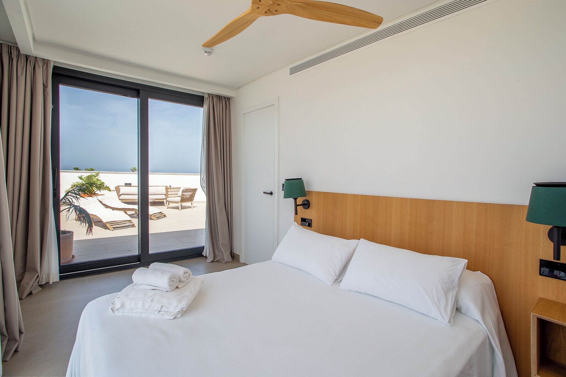 A hotel room with a bed, a ceiling fan, a nightstand, a lamp, and a view of the beach through a sliding door.