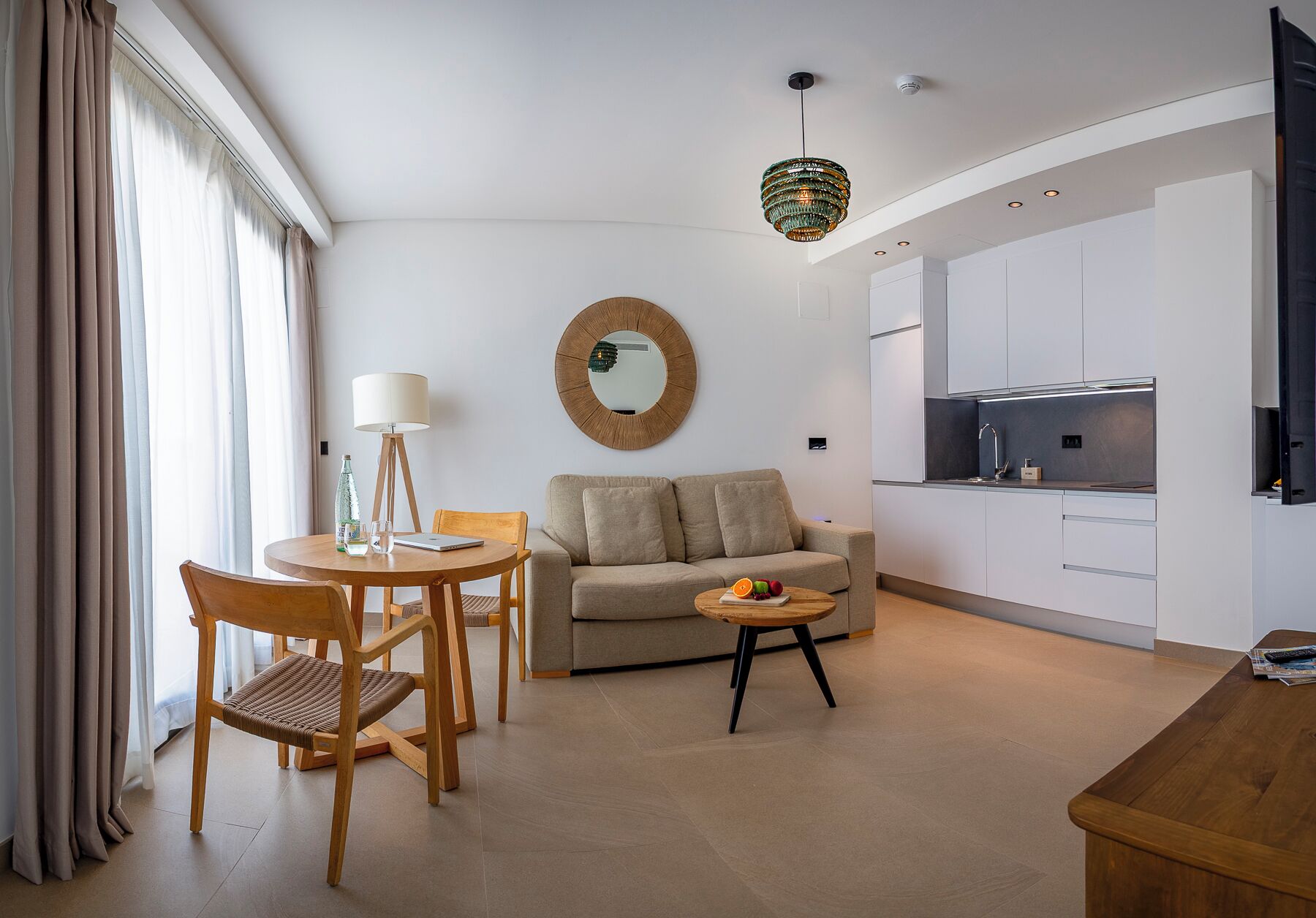A modern living room with a sofa, a dining table with chairs, a small round table with a lamp, and a kitchen area with cabinets and a sink.