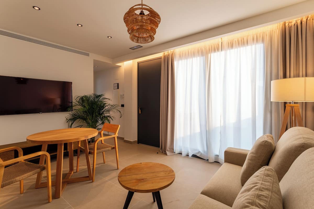 A modern living room with a sofa, a dining table with chairs, a TV mounted on the wall, and a large window with curtains.