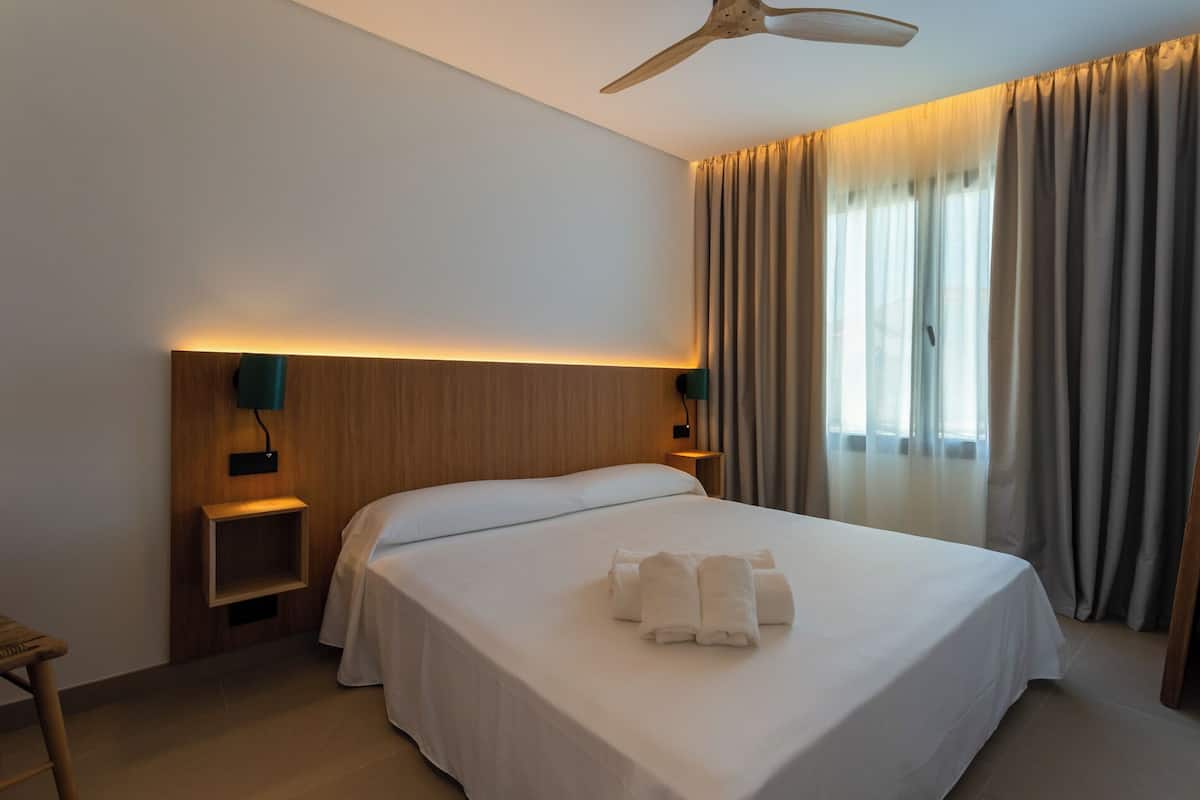 A hotel room with a large bed, two bedside tables, a ceiling fan, and a window with curtains.