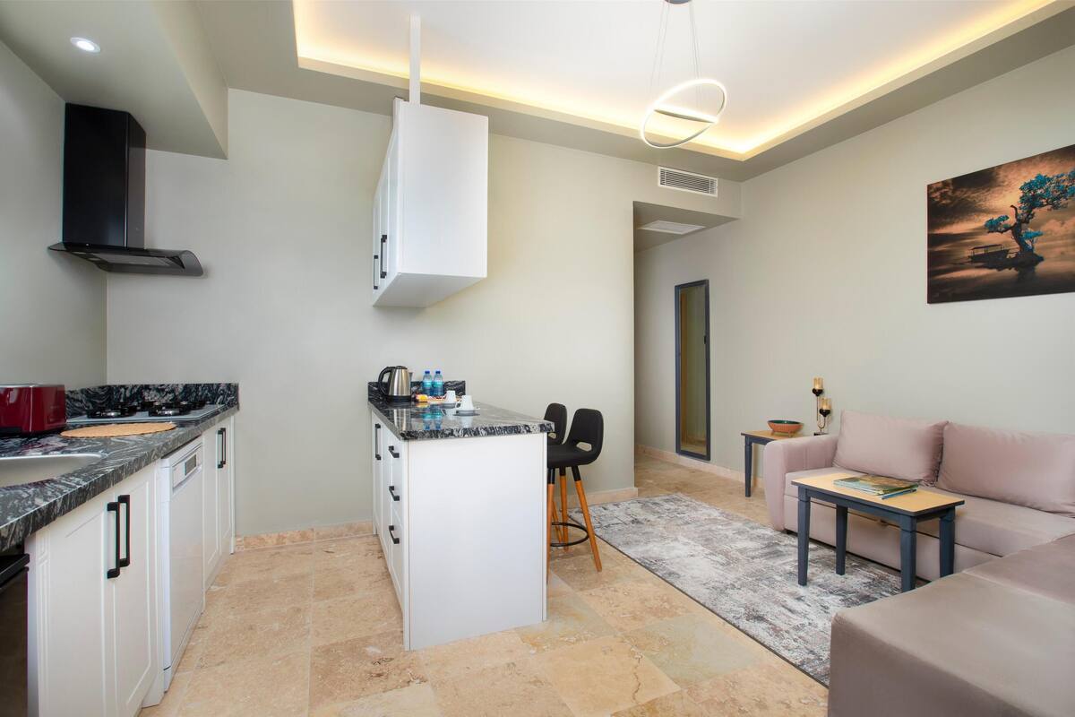 A modern kitchen with white cabinets, a marble countertop, and a built-in oven. There is a small island with bar stools and a painting on the wall.