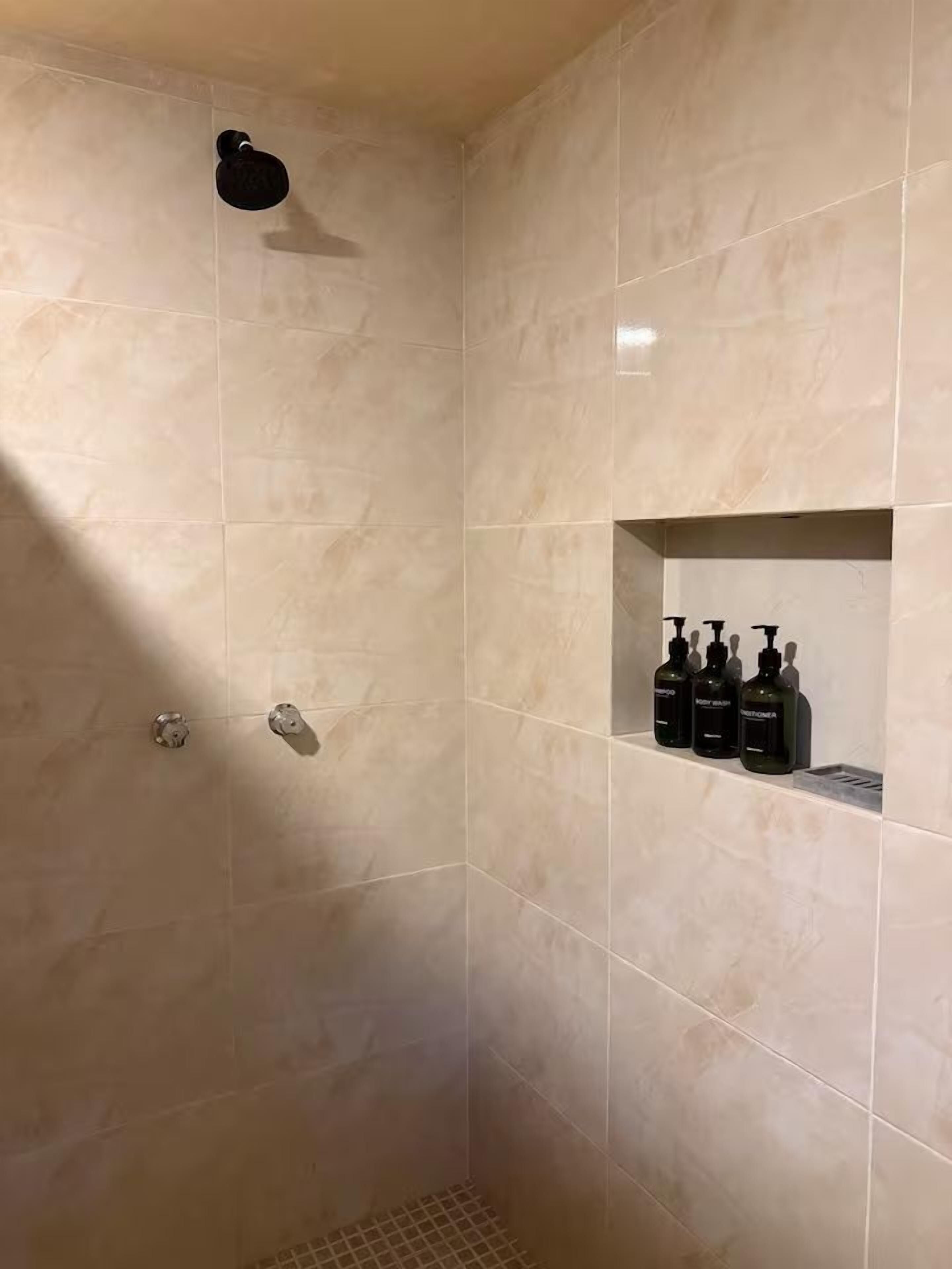 A shower area with beige tiles, a showerhead, and a shelf with three bottles.