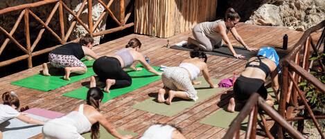 Een groep mensen doet yoga op een houten terras met een rustieke houten reling en een stenen muur op de achtergrond.