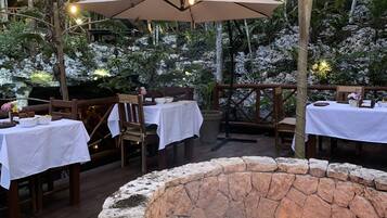 An outdoor dining area with stone fire pit, wooden tables, chairs, and string lights.