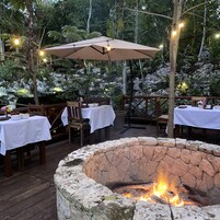 Un espacio exterior para comer con fogón de piedra, mesas de madera, sillas y luces colgantes.
