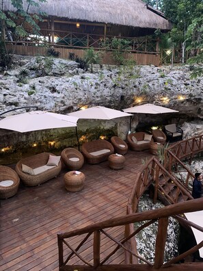 Une terrasse en bois, agrémentée de meubles en osier et de parasols, entourée d’un jardin de rochers naturels et d’une structure à toit de chaume.