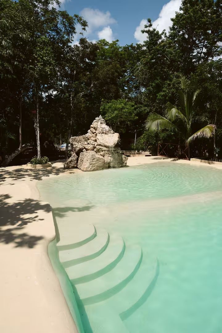 Una piscina cristalina con una formación rocosa y palmeras al fondo.