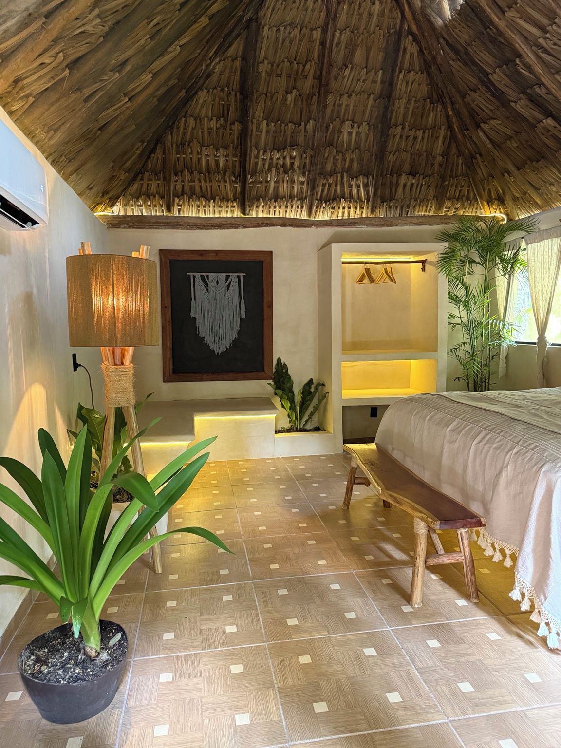 A room with a thatched ceiling, a bed, a wall-mounted lamp, a framed macrame piece, and a potted plant.