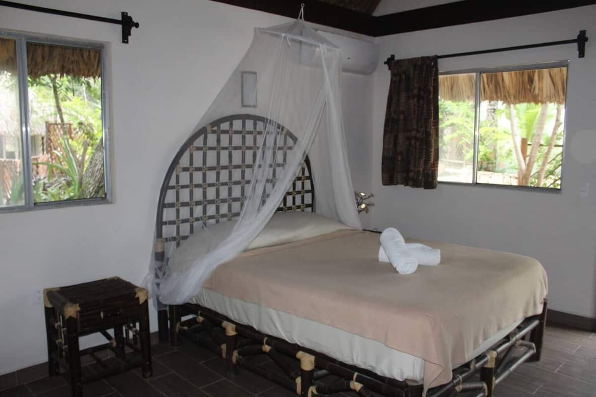 A single bed with a canopy, a small wooden stool, and a window with curtains.