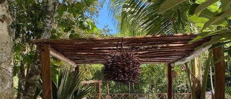 A wooden pergola with a hanging decoration, a pool, and surrounding tropical vegetation.