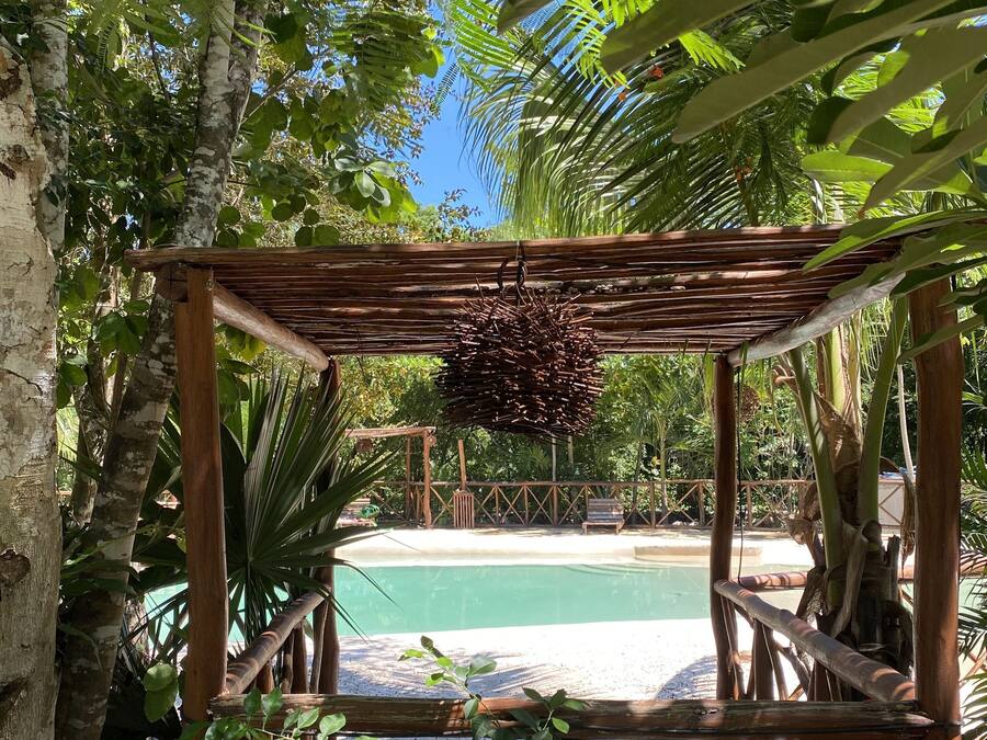 A wooden pergola with a hanging decoration, a pool, and surrounding tropical vegetation.