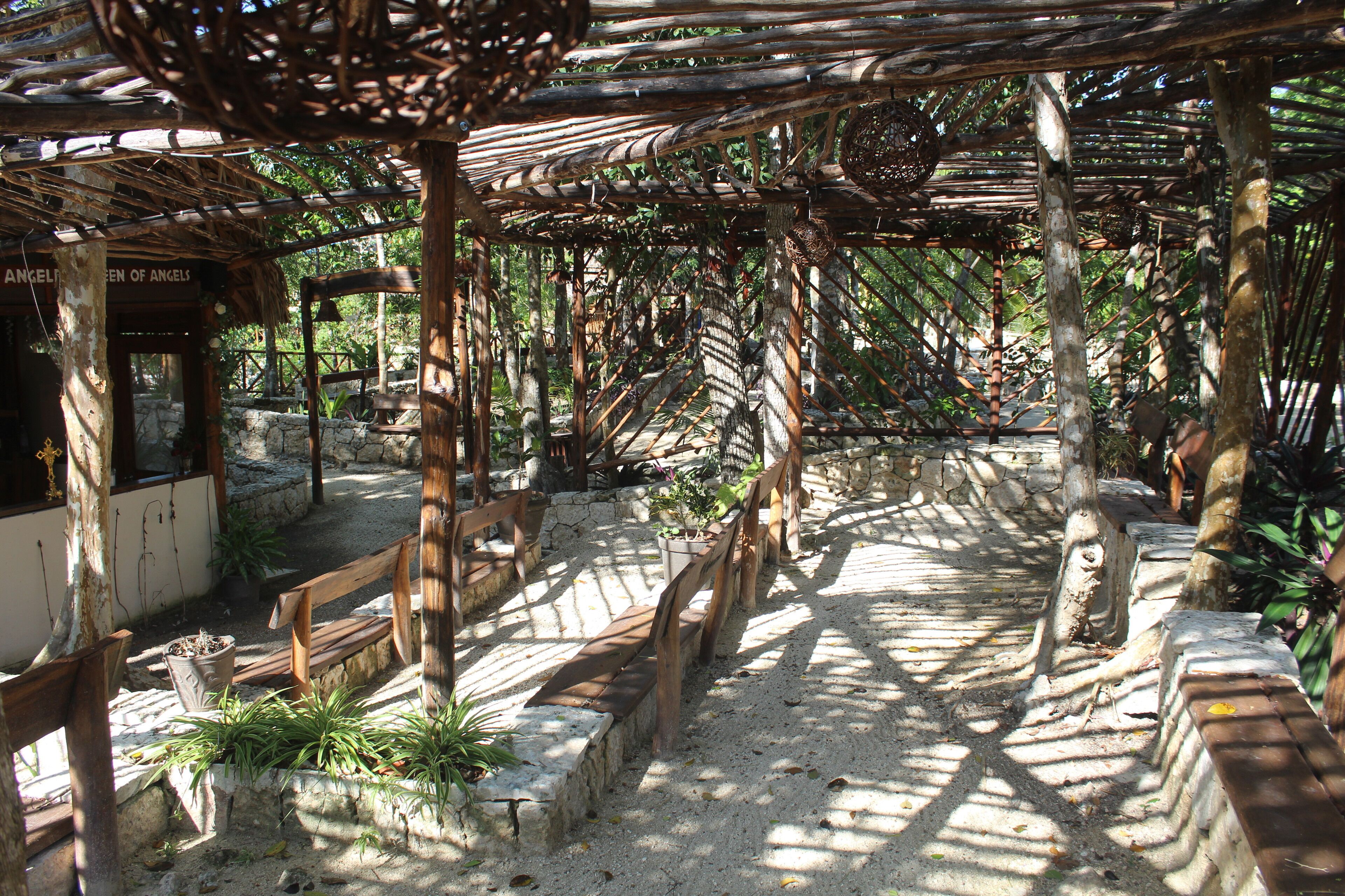 A wooden pergola structure with benches and potted plants, surrounded by stone walls and greenery.