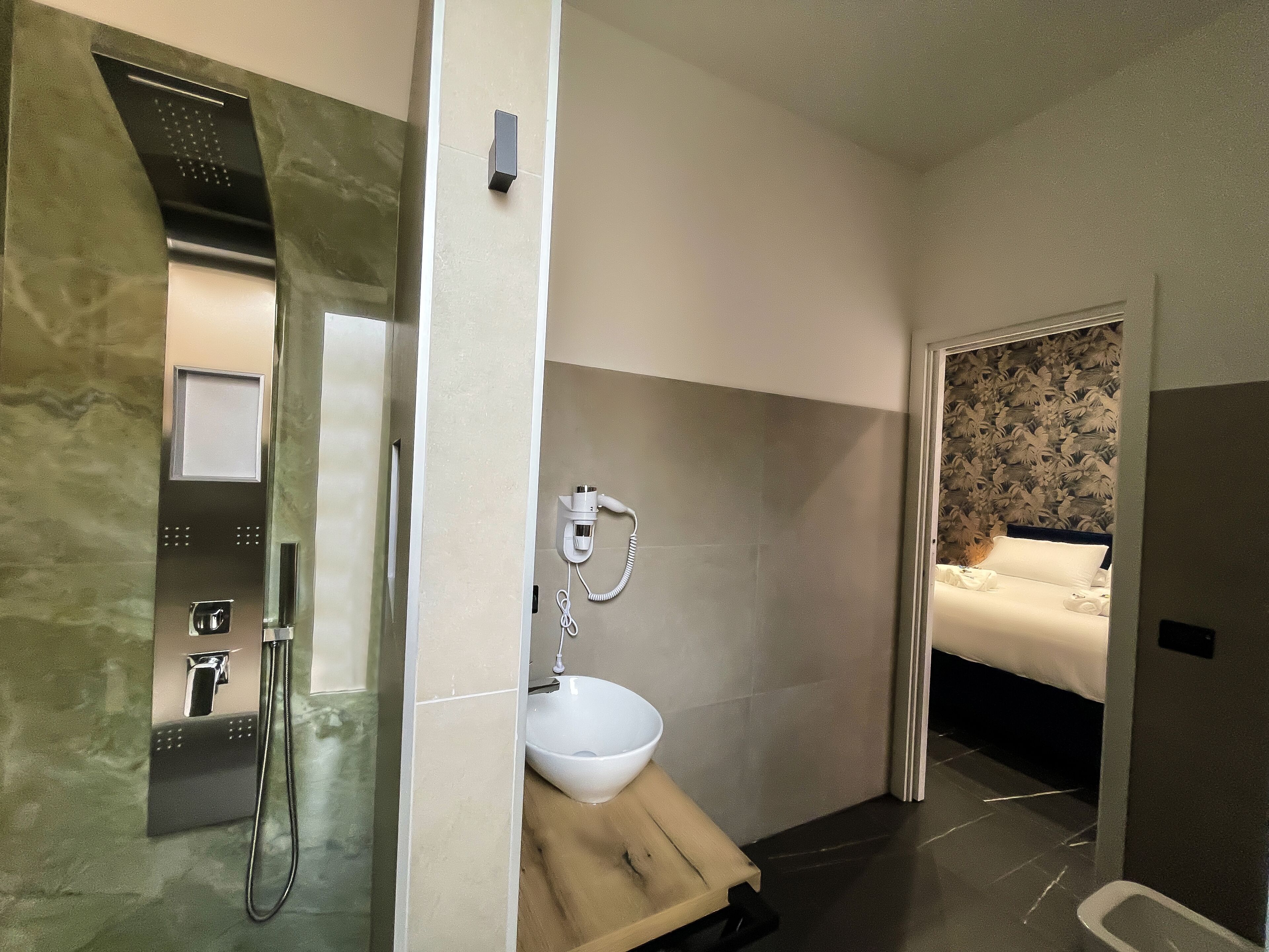 A modern bathroom with a glass-enclosed shower, a white sink, and a bed with a floral headboard in the adjacent room.