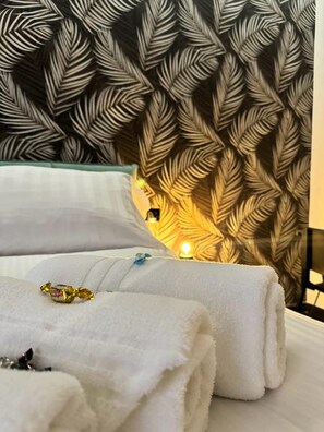 Uma cama bem arrumada com toalhas brancas e um travesseiro decorativo, uma parede com papel de parede estampado com folhas, e uma pequena luminária de cabeceira.