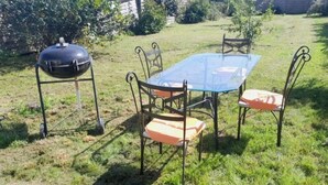 An outdoor dining area with a glass table and metal chairs, a barbecue grill, and a grassy lawn. - Location Maison 3 Chambres Circuit des 24H (Le Mans)