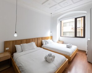 A hotel room with two beds, wooden headboards, white bedding, a wooden nightstand, a bedside table, a window with a view of buildings, and a ceiling with a decorative pattern.