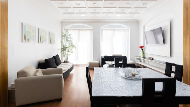 A modern living room with a sofa, dining table, and chairs. There are two windows with white frames and a ceiling with decorative panels. - Ste. Presidencial • Steps from the Catedral (Luis Cordero Cuenca)