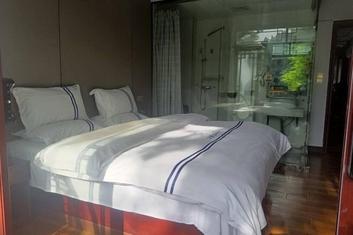 A modern hotel room with a large bed, a glass shower enclosure, and a view of the city.
