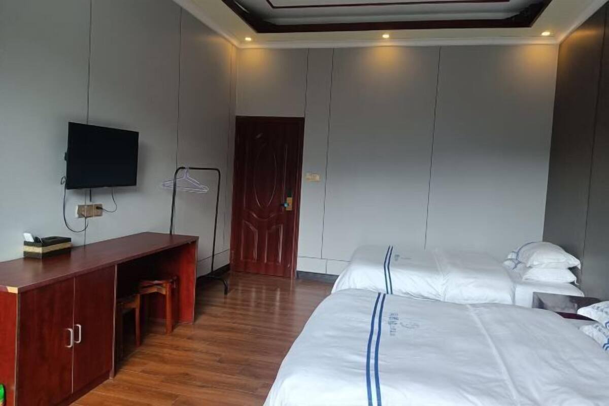 A hotel room with a wooden desk, a television, a wardrobe, a bed with white bedding, and a door.