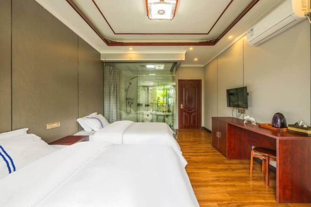 A hotel room with two beds, a desk, a television, and a bathroom visible through a glass door.