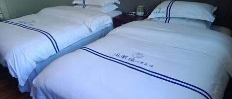 Habitación de hotel con dos camas, cada una con ropa blanca y detalles azules. Hay una mesita de noche con una lámpara y una botella de agua.