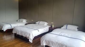 Habitación de hotel con cuatro camas individuales, cada una con ropa blanca y cabeceros oscuros. El suelo es de madera y las paredes son de un tono neutro.