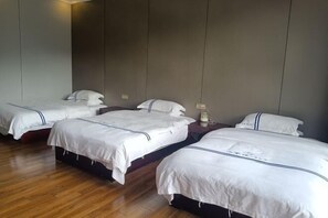 Une chambre d’hôtel avec quatre lits simples, chacun doté de draps blancs et d’une tête de lit sombre. La chambre est dotée d’un plancher en bois et d’un mur aux couleurs neutres.