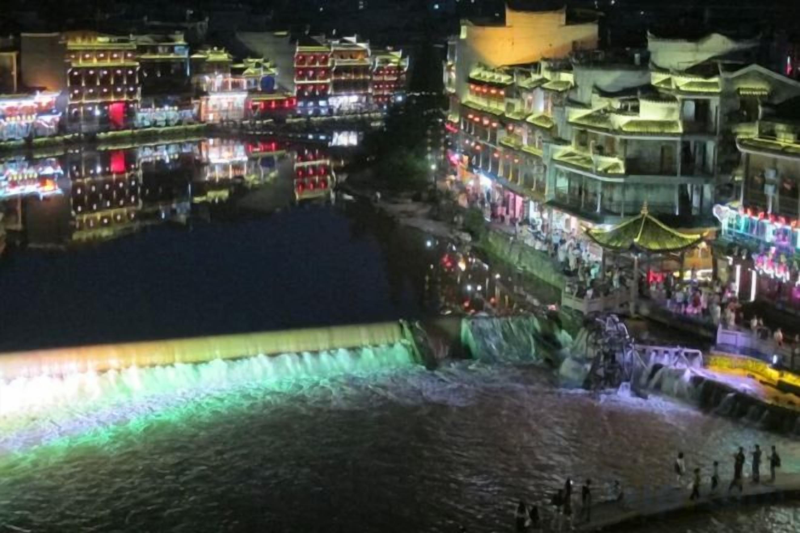 A riverside view of a traditional town with buildings along the water, a waterfall, and people walking along the riverbank.