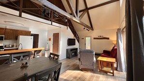 Un vaste espace de vie doté d’un haut plafond, de poutres apparentes, d’une table à manger avec des chaises, d’un coin salon avec un canapé et un fauteuil, et d’un téléviseur fixé au mur.